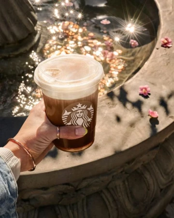 Hand holding a Starbucks drink near a fountain with flower petals, creating a refreshing and visually appealing beverage presentation.