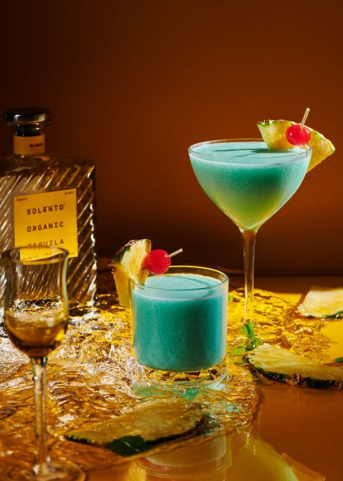 Two turquoise cocktails garnished with pineapple and cherries are served on ice beside a bottle of organic tequila, studio lighting, rich color.