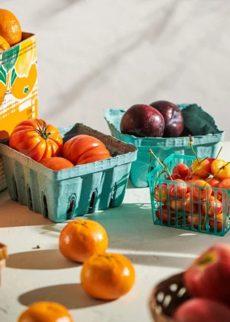 Still life of fresh fruits including tomatoes, plums, cherries, and oranges arranged in colorful baskets on a white surface.
