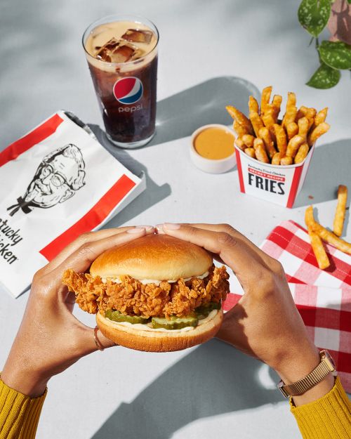 Overhead shot of a fried chicken sandwich held by a woman's hands, with fries, sauce, and a Pepsi in the background.