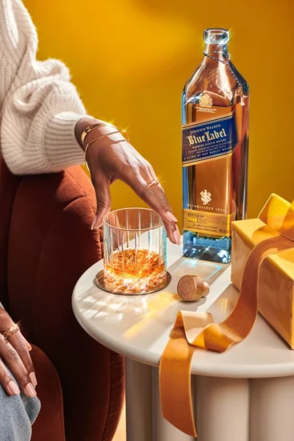 Person pours Johnnie Walker Blue Label whiskey into a glass. Bottle, gift box, and ribbon sit on a white round table against a yellow background.