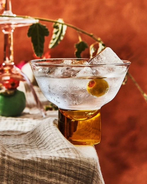 Close-up of a clear martini glass filled with ice and a green olive, standing on a grey linen fabric against an orange backdrop.