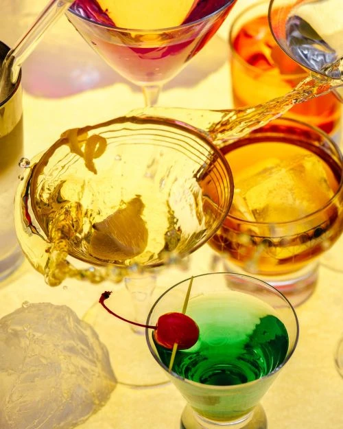 Assorted cocktails in stemmed glasses with ice and garnishes, displaying a variety of colors and ingredients on a light surface.