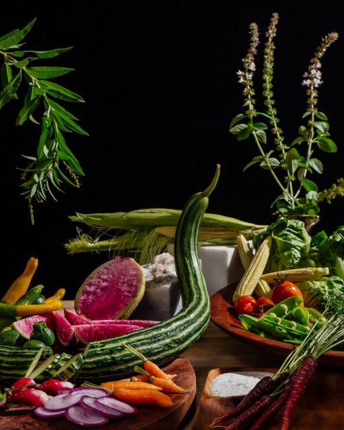 Still life food photography featuring an assortment of colorful, fresh vegetables including corn, carrots, radishes, and leafy greens.