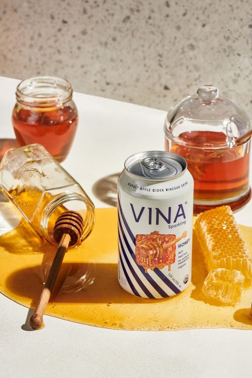 Still life of a can of Vina Sparkling Apple Cider Vinegar drink, honeycomb, honey dipper, and jars of honey on a white surface.