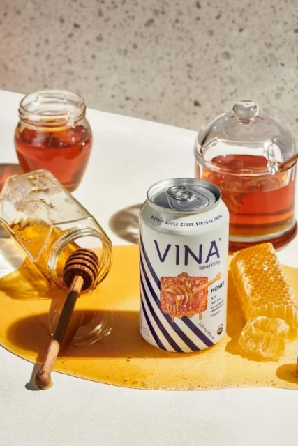 Still life of a can of Vina Sparkling Apple Cider Vinegar drink, honeycomb, honey dipper, and jars of honey on a white surface.