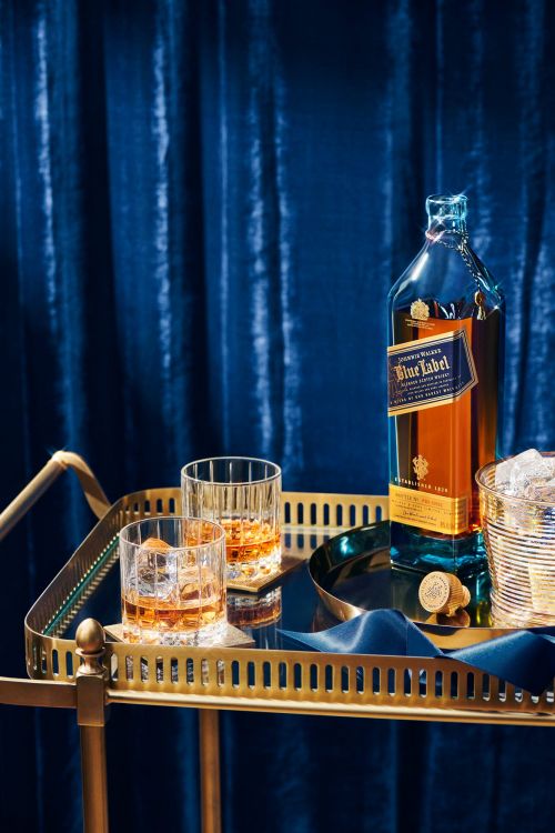 Bottle of Johnnie Walker Blue Label and two glasses with ice on a gold-colored bar cart with a blue velvet curtain backdrop.