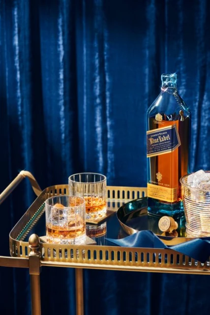Bottle of Johnnie Walker Blue Label and two glasses with ice on a gold-colored bar cart with a blue velvet curtain backdrop.