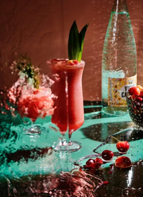 A vibrant red cocktail is garnished with pineapple fronds and surrounded by cherries, set on a glossy table with water droplets.