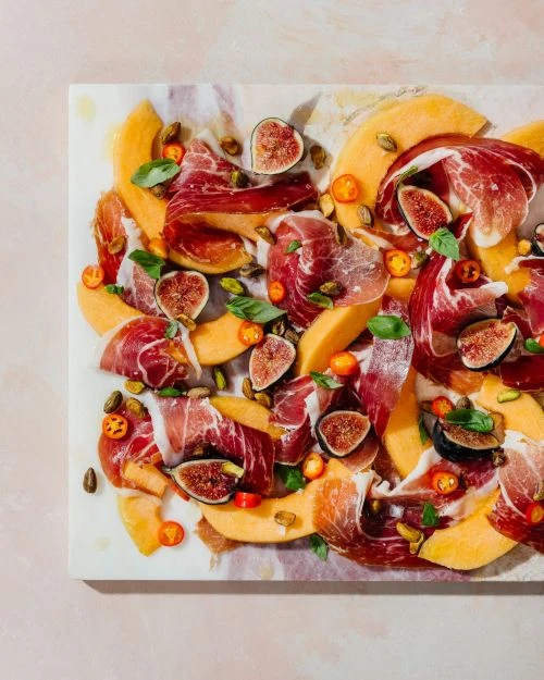 Overhead view of sliced cantaloupe, figs, and prosciutto arranged on a marble serving board. Food photography for catering or recipe concept.