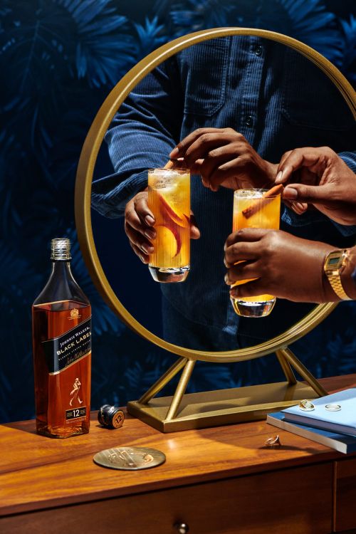 Person putting a cinnamon stick in a glass of amber liquid. A bottle of Johnnie Walker whiskey is on the wooden table.