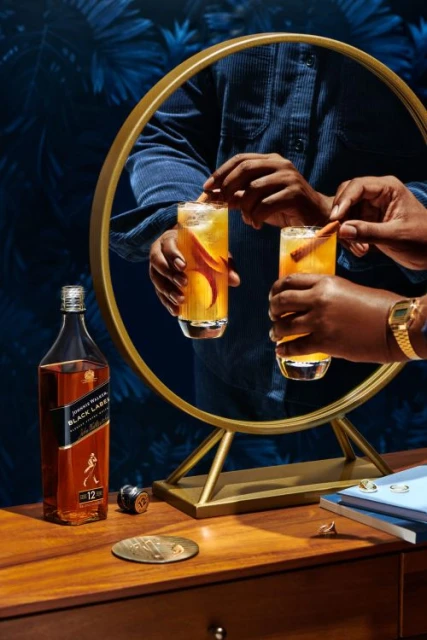 Person putting a cinnamon stick in a glass of amber liquid. A bottle of Johnnie Walker whiskey is on the wooden table.