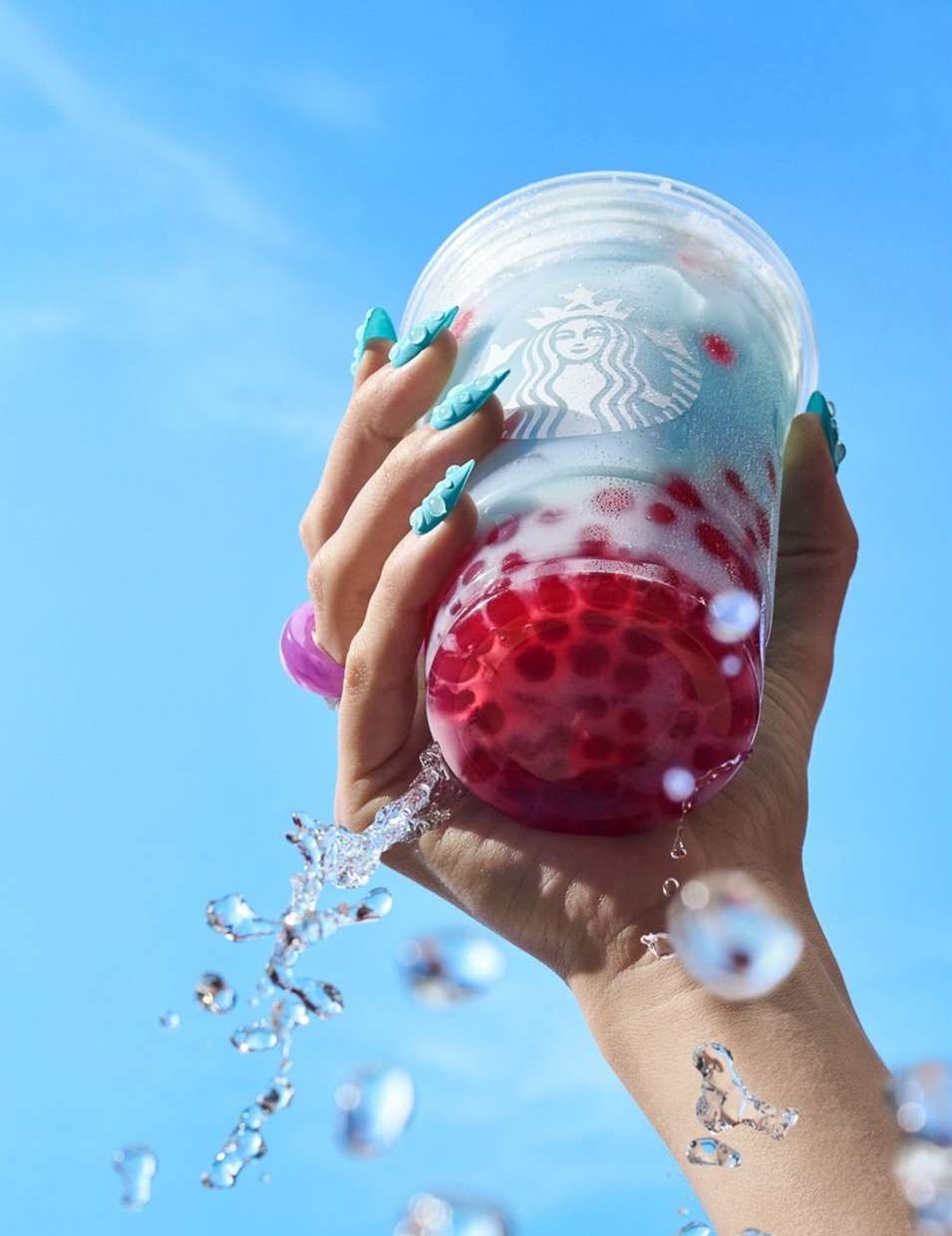 Hand holding a Starbucks drink with red boba pearls and blue liquid, water splashing against a blue sky background in a bright, clean setting.