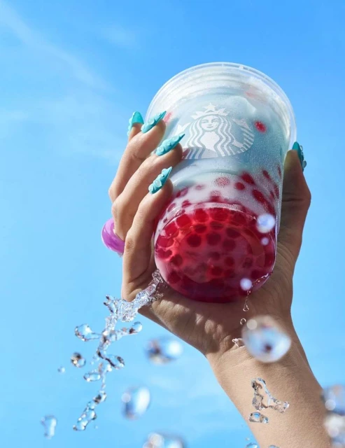 Hand holding a Starbucks drink with red boba pearls and blue liquid, water splashing against a blue sky background in a bright, clean setting.