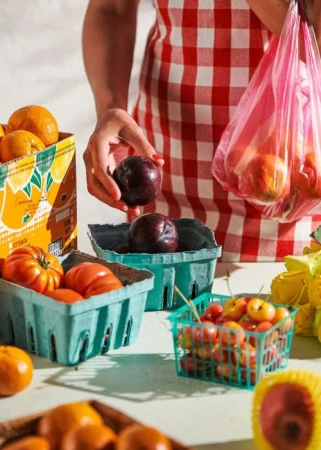 Overhead shot of person in gingham apron holding a plum. Fruit in baskets and a plastic bag with peaches on a white table.