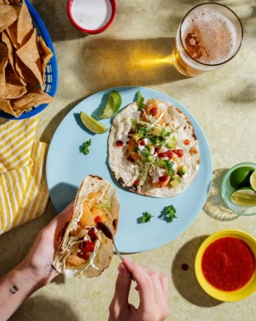 Overhead shot of hands preparing shrimp tacos on a blue plate, served with tortilla chips, beer, and lime wedges. Food photography.