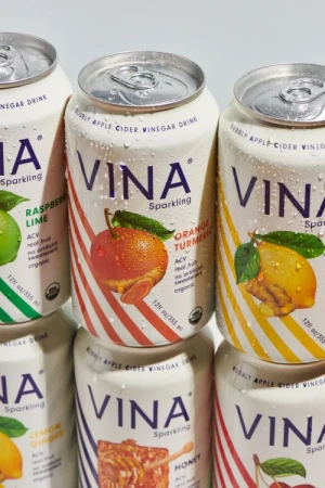 Several cans of Vina sparkling apple cider vinegar drink are stacked together, showcasing different fruit flavors on white background.