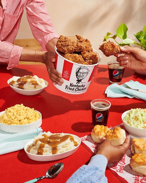 Overhead shot of fast-food fried chicken, biscuits, and side dishes served at a red table with several people sharing the meal.