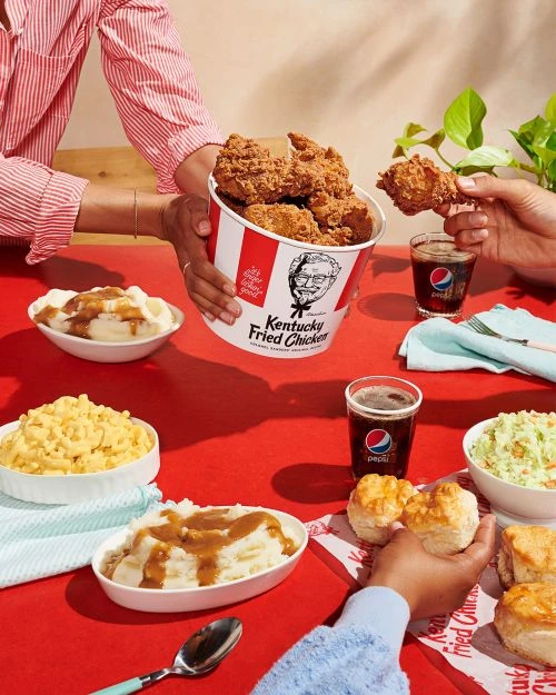 Overhead shot of fast-food fried chicken, biscuits, and side dishes served at a red table with several people sharing the meal.