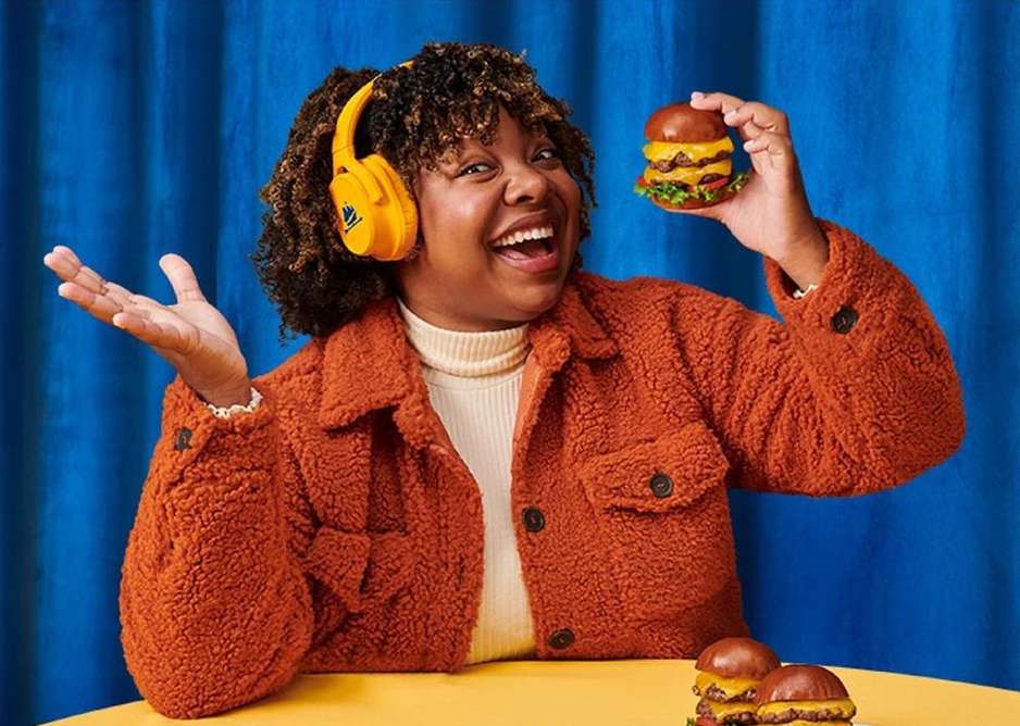Cheerful woman with headphones smiles while holding a double cheeseburger against a blue backdrop, two additional burgers sit on a table.