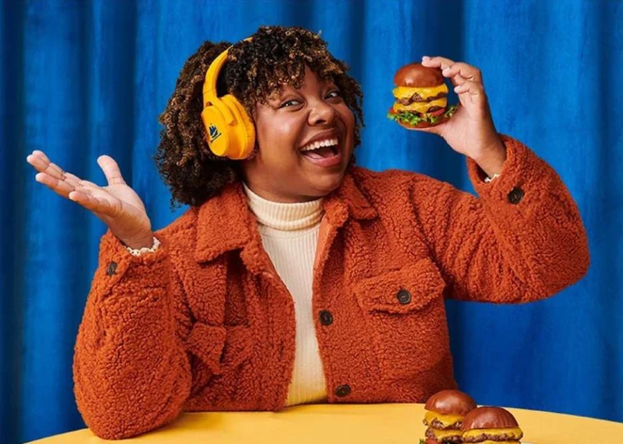 Cheerful woman with headphones smiles while holding a double cheeseburger against a blue backdrop, two additional burgers sit on a table.