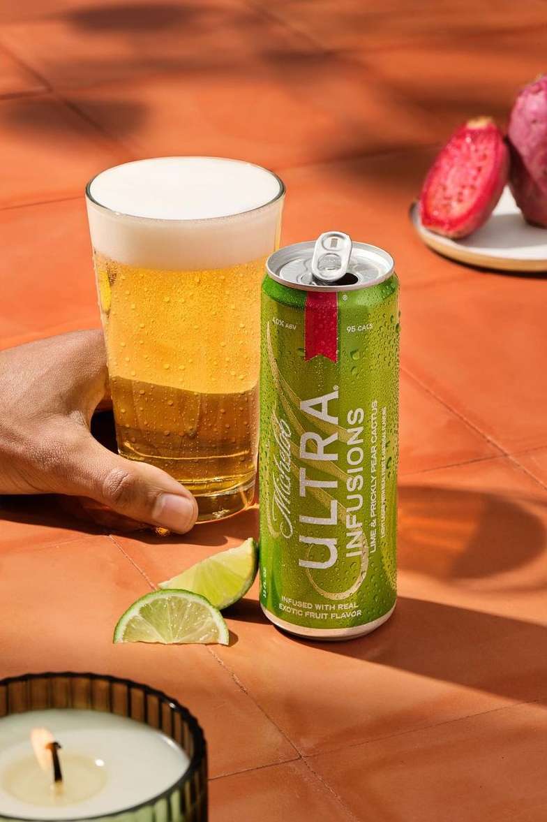 Michelob Ultra Infusions beer can and glass of beer with lime slices and cactus fruit on an orange tiled surface, outdoor setting.