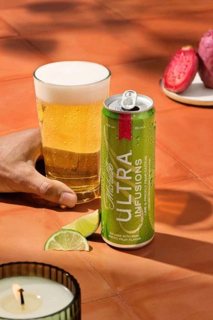 Michelob Ultra Infusions beer can and glass of beer with lime slices and cactus fruit on an orange tiled surface, outdoor setting.