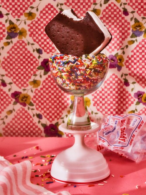 An ice cream sandwich, with a bite taken out, sits atop a glass filled with colorful sprinkles, creating a playful food photography.