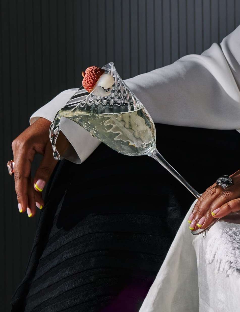 Woman in formal wear holds a sparkling wine glass with a lychee. The drink is chilled; the glass is fluted, and the background is grey.