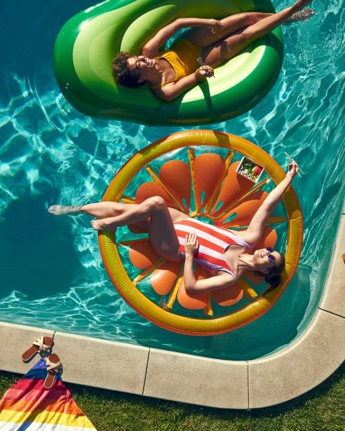 Two women relax on fruit-shaped inflatables in a blue swimming pool on a sunny day, with a rainbow towel and sandals nearby.