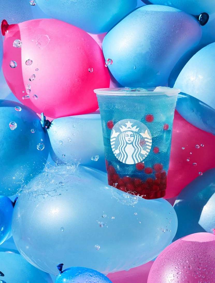 Starbucks drink with boba sits on a blue balloon surrounded by pink and blue balloons and water droplets in an eye-level studio shot.