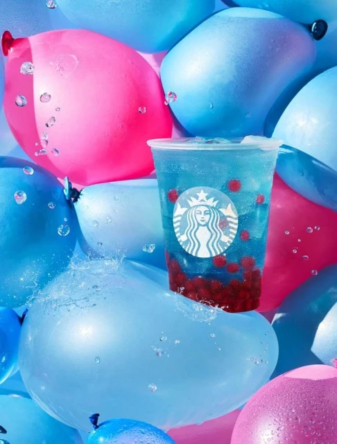 Starbucks drink with boba sits on a blue balloon surrounded by pink and blue balloons and water droplets in an eye-level studio shot.