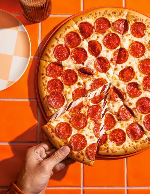 Overhead shot of a hand grabbing a slice of hot, cheesy, pepperoni pizza from a whole pizza on a orange tiled surface with a plate.