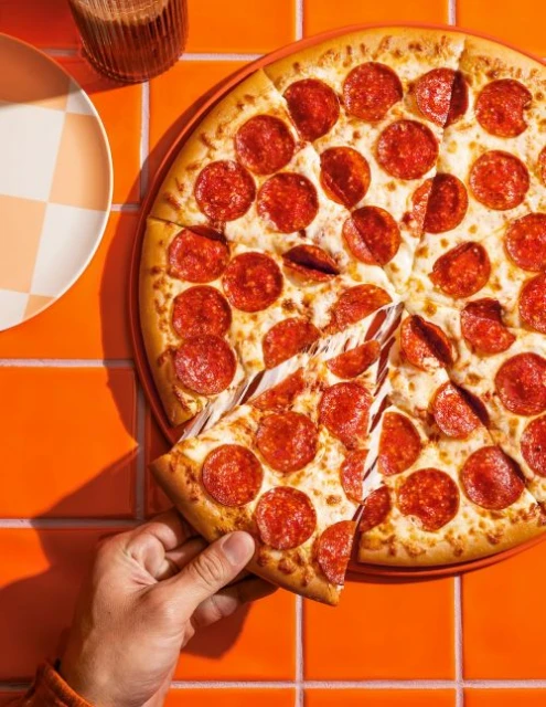 Overhead shot of a hand grabbing a slice of hot, cheesy, pepperoni pizza from a whole pizza on a orange tiled surface with a plate.