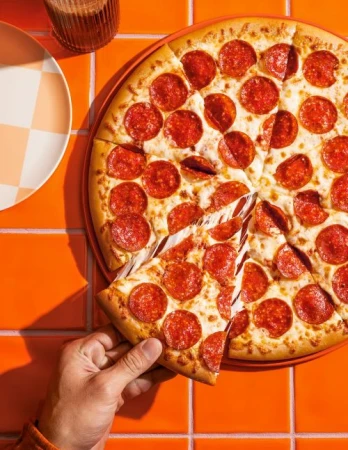 Overhead shot of a hand grabbing a slice of hot, cheesy, pepperoni pizza from a whole pizza on a orange tiled surface with a plate.