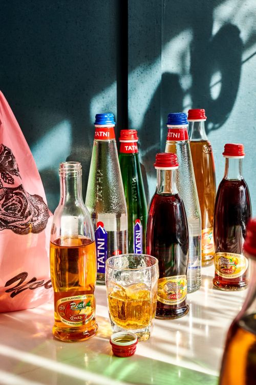 Still life photograph featuring clear glass bottles filled with colorful beverages and a glass with ice on a white table, shadowed by a blue wall.