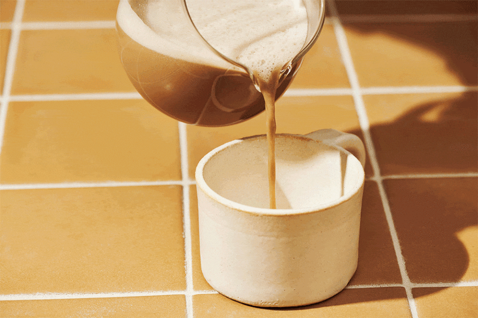 Foamy brown liquid, possibly coffee or tea, is being poured from a clear glass pitcher into a white ceramic mug on a tan tiled surface.