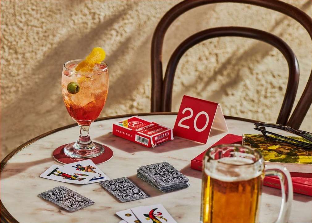 Cocktail and playing cards are displayed on a marble table next to a glass beer mug, Modiano cards, and a red table number 20.