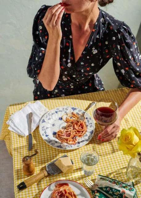 Woman in a floral dress enjoys spaghetti at a table set with a yellow checkered tablecloth, glass of wine, and dinnerware.