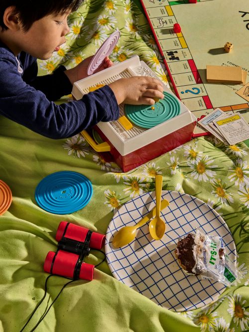 Child playing with a vintage toy record player on a daisy-patterned blanket alongside a Monopoly game, plate with dessert, and binoculars.