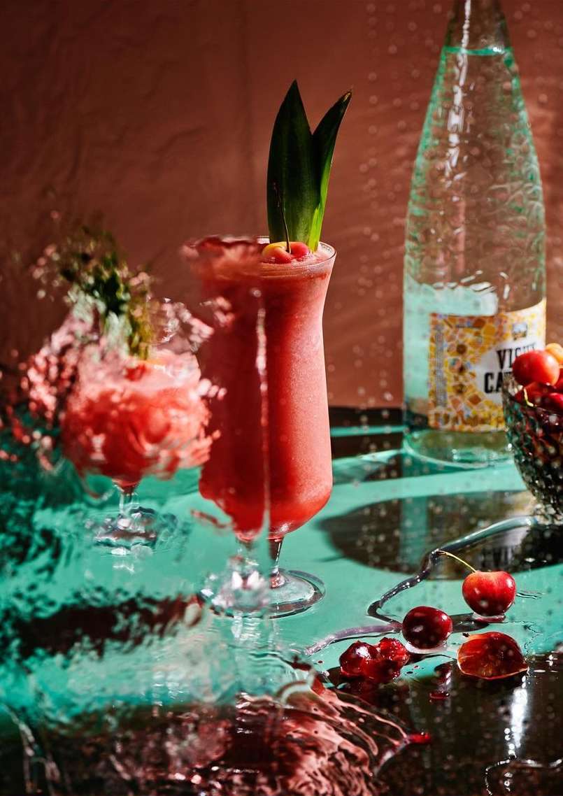 Two glasses of red cocktails, garnished with cherries and pineapple leaves, sit on a wet surface next to a bottle of sparkling water.