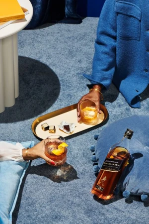 Two people hold cocktails near a bottle of Johnnie Walker Black Label whisky on a blue textured surface, accompanied by wrapped chocolates and a small table.