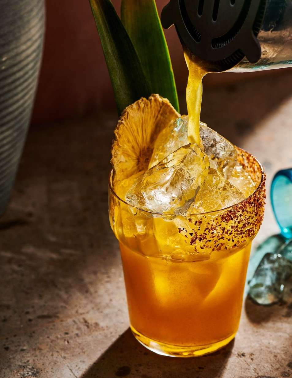 Cocktail pouring from a strainer into a glass filled with ice, garnished with pineapple leaves and slice, lit with studio lighting.