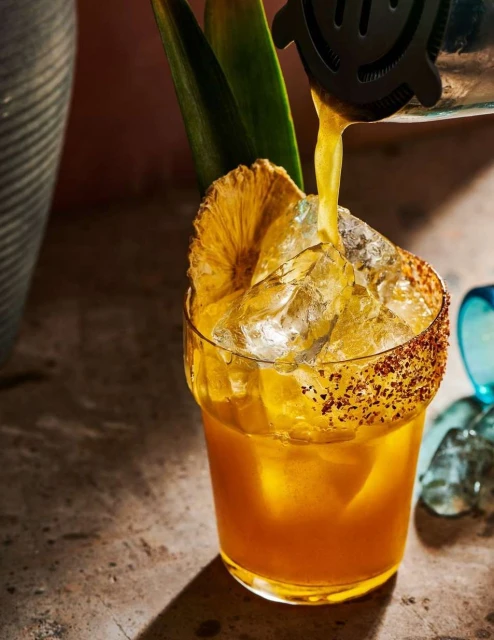 Cocktail pouring from a strainer into a glass filled with ice, garnished with pineapple leaves and slice, lit with studio lighting.
