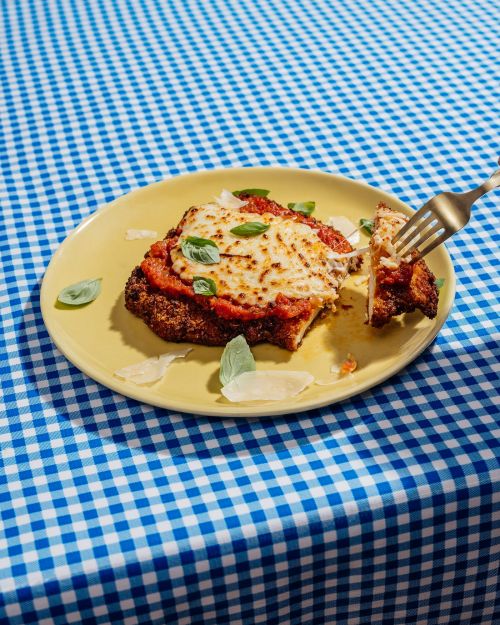 Overhead shot of chicken parmesan on a plate with a fork, tomato sauce, cheese, and basil, set on a blue and white checkered tablecloth.