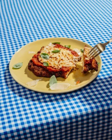 Overhead shot of chicken parmesan on a plate with a fork, tomato sauce, cheese, and basil, set on a blue and white checkered tablecloth.