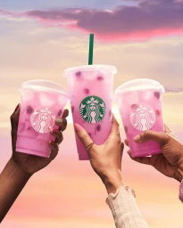 Three hands holding clear plastic cups of pink iced beverages with green straws against a sunset sky background
