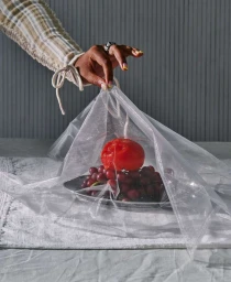 Hand in patterned sleeve lifting sheer mesh fabric over a metal tray of red grapes and a single tomato