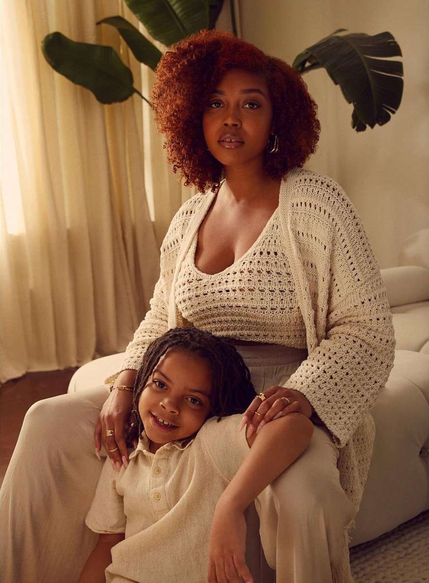 Portrait of an African-American mother and child in a light-filled indoor setting, both wearing matching knitwear in neutral tones, smiling.