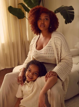 Portrait of an African-American mother and child in a light-filled indoor setting, both wearing matching knitwear in neutral tones, smiling.